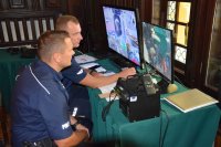Na fotografii widać dwóch policjantów obserwujących monitory, na których widać uczestników imprezy.