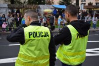 Na fotografii widać odwróconych tyłem policjantów. W tle fotografii widać stojący przy ulicy tłum ludzi.