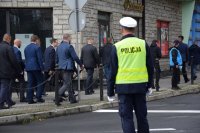 Na fotografii widać odwróconego tyłem policjanta. W tle fotografii widać przemieszczających się ulicą parlamentarzystów.