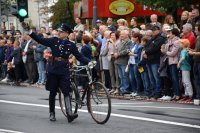 Na fotografii widać mężczyznę, który idzie ulicą i prowadzi rower. Ubrany jest w stary policyjny mundur. Po obydwu stronach ulicy znajduje się tłum ludzi.