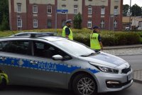 Na fotografii widać rondo, na którym znajduje się policyjny radiowóz.  Obok samochodu  stoi policjant i policjantka.
