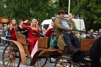 Na fotografii widać jadących karocą uczestników pochodu. Przebrani są za historyczne postacie. Po obydwu stronach ulicy znajduje się tłum ludzi.