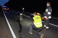 Na fotografii widać drogę, na której znajduje się rozbity motocykl. Przed motocyklem, na drodze klęczy policjant, który wykonuje zdjęcie jednośladu. Obok policjanta stoi strażak. W tle fotografii widać wóz strażacki.