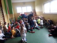 Na fotografii  grupę siedzących na podłodze dzieci. Wśród nich widać policjanta, który rozdaje dzieciom odblaskowe zawieszki.