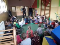 Na fotografii  widać grupę siedzących na podłodze dzieci. Przed nimi stoją dwaj policjanci.