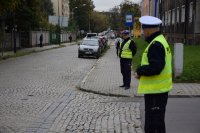 Na fotografii widać ulicę, przy której stoi dwóch policjantów. Wzdłuż ulicy zaparkowanych jest kilkanaście samochodów.
