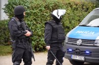 Na fotografii widać dwóch policjantów stojących obok radiowozu. Mundurowi ubrani są w kamizelki przeciwuderzeniowe  i hełmy. Jeden z nich w ręku trzyma pałkę, a drugi broń gładkolufową.