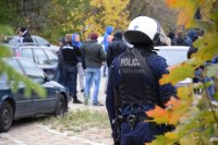 Na fotografii widać odwróconego tyłem policjanta ubranego w kamizelkę i hełm. W tle fotografii widać zaparkowane samochody i stojące obok nich osoby.