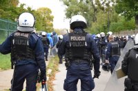 Na fotografii widać odwróconych tyłem policjantów, którzy są ubrani w kamizelki i hełmy. W tle fotografii widać innych policjantów i grupę osób.