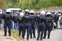 Na fotografii widać kilku odwróconych tyłem policjantów, którzy są ubrani w kamizelki i hełmy. Mundurowi stoją obok grupy osób.