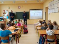 Na fotografii widać szkolną klasę. W szkolnych ławkach siedzi grupa dzieci, obok tablicy stoi dwóch policjantów.