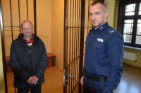 Na fotografii widać policjanta, który otwiera drzwi do policyjnej celi. Obok policjanta, przy kracie stoi mężczyzna.