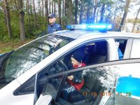 Na fotografii widać policyjny radiowóz. W środku za kierownicą siedzi dziewczynka, a obok stoi policjant.