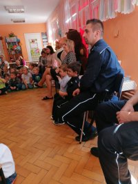 Na fotografii widać klasę, w której siedzi grupa osób. Wśród nich znajdują się policjanci oraz przedszkolaki.