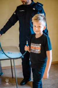 Na fotografii widać Miłosza i policjanta. Chłopiec stoi w pomieszczeniu i trzyma w dłoni ustnik, przez który do urządzenia wdmuchuje się powietrze podczas badania trzeźwości.