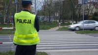 Na fotografii widać przejście dla pieszych. Obok niego stoi odwrócony tyłem policjant. Ma na sobie odblaskową kamizelkę z napisem POLICJA. W tle fotografii widać przejeżdżający samochód.