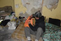 Na fotografii widać pomieszczenie, w którym porozrzucane są różne śmieci. Na podłodze rozłożony jest materac, na którym siedzi mężczyzna i wkłada coś do plecaka.