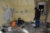 Na fotografii widać pomieszczenie, w którym porozrzucane są różne śmieci. Na podłodze rozłożony jest materac, na którym siedzi mężczyzna. Przed siedzącym mężczyzną stoi pracownik noclegowni
