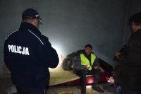 Na fotografii widać pomieszczenie, w którym na kanapie siedzi mężczyzna ubrany w odblaskową kamizelkę. Z lewej strony zdjęcia widać odwróconego tyłem policjanta