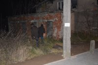 Na fotografii widać dwóch, odwróconych tyłem mężczyzn. Stoją w zaroślach i patrzą na opuszczony budynek