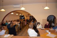 Na fotografii widać siedzące przy stołach osoby. W tle zdjęcia widać policjantów, jeden z nich trzyma w ręku mikrofon