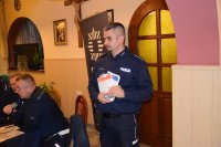 Na fotografii widać policjantów. Dwóch z nich siedzi przy stole, a trzeci stoi i trzyma w ręku ulotki.