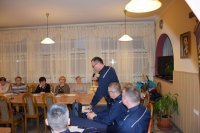 Na fotografii widać siedzące przy stołach osoby. Na zdjęciu widać również policjantów, jeden z nich trzyma w ręku mikrofon