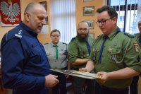 Na fotografii widać od lewej Komendanta Powiatowego Policji w Tarnowskich Górach insp. Krzysztofa Hajdę oraz harcerza, który przekazuje komendantowi dyplom z podziękowaniem za współpracę. w tle fotografii widać pozostałych harcerzy.