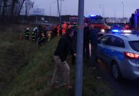 Na fotografii widać pobocze drogi, rów, w którym leży na boku samochód osobowy. Obok samochodu znajduje się kilku strażaków. Na drodze widać policyjny radiowóz oraz wozy strażackie.