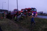 Na fotografii widać pobocze drogi, rów, w którym leży na boku samochód osobowy. Obok samochodu znajduje się kilku strażaków. Na drodze widać  wozy strażackie.