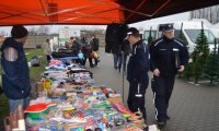 Z lewej strony fotografii widać stragany. Na stołach porozkładane są różne przedmioty. Obok stołu stoi mężczyzna. Z prawej strony zdjęcia widać dwóch policjantów oraz innych ludzi oglądających towar znajdujący się na stołach.