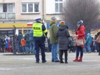 Na fotografii od lewej strony widać policjanta oraz stojące obok niego trzy osoby. W le zdjęcia widać tłum ludzi.
