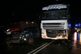 Na fotografii widać uszkodzony samochód ciężarowy, obok którego widać rozbity przód samochodu osobowego, W tle fotografii widać policyjny radiowóz