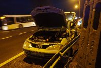 Na fotografii widać rozbity samochód osobowy. Pojazd ma podniesioną do góry maskę oraz tylną klapę, a bok od strony kierowcy jest zgnieciony. W tle zdjęcia widać przejeżdżający autobus.
