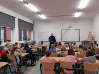 Na fotografii widać szkolną klasę, w której przy stolikach, siedzi duża grupa dzieci. Pod tablicą widać policjanta z wydziału ruchu drogowego