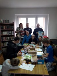 Na fotografii widać pomieszczenie biblioteki, w którym przy stole siedzi grupa dzieci. Przy oknie, stoją dwaj policjanci oraz kobieta i mężczyzna. Policjanci trzymają w rękach plakaty z nazwami kampanii edukacyjno-profilaktycznych