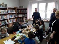 Na fotografii widać pomieszczenie biblioteki. Przy stoliku siedzą dzieci, a obok nich stoją dwaj policjanci i kobieta. Za dziećmi widać regały, na których znajdują się książki