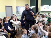 Na fotografii widać szkolną klasę, w której przy stolikach, siedzi duża grupa dzieci. Wśród nich znajdują się policjant, który rozdaje dzieciom ulotki