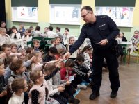 Na fotografii widać szkolną klasę, w której przy stolikach, siedzi duża grupa dzieci. Wśród nich znajdują się policjant, który rozdaje dzieciom ulotki
