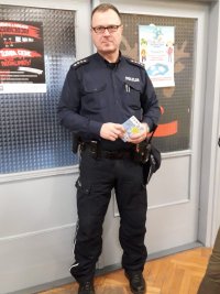 Na fotografii widać szkolną klasę, w której przy drzwiach stoi policjant. Na drzwiach przyklejone są plakaty kampanii edukacyjno- profilaktycznych.