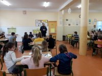 Na fotografii widać szkolną klasę, w której przy stolikach, siedzi duża grupa dzieci. Wśród nich znajdują się policjant