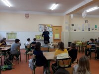 Na fotografii widać szkolną klasę, w której przy stolikach, siedzi duża grupa dzieci. Wśród nich znajdują się policjant, który w dłoni trzyma plakat kampanii