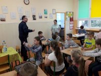 Na fotografii widać szkolną klasę, w której przy stolikach, siedzi duża grupa dzieci. Wśród nich znajdują się policjant, który rozdaje dzieciom ulotki