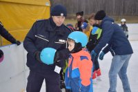 Na fotografii widać lodowisko, na którym znajdują się policjant i grupa młodych łyżwiarzy. Policjant trzyma w ręku niebieski kask narciarski