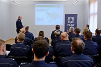 grupa policjantów siedzących na krzesłach. Przed rzutnikiem stoi  Komendant Powiatowy Policji w Tarnowskich Górach insp. Krzysztof Hajda