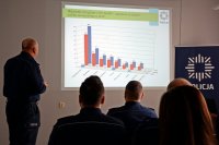 grupa policjantów siedzących na krzesłach. Przed rzutnikiem stoi  Komendant Powiatowy Policji w Tarnowskich Górach insp. Krzysztof Hajda