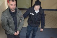 policjant prowadzi zatrzymanego mężczyznę, który na rękach ma zapięte kajdanki. W tle widać zaparkowane samochody