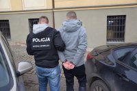 policjant prowadzi zatrzymanego mężczyznę, który na rękach ma zapięte kajdanki. W tle widać zaparkowane samochody