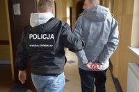 policjant prowadzi zatrzymanego mężczyznę, który na rękach ma zapięte kajdanki. W tle widać zaparkowane samochody