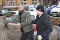 policjant prowadzi zatrzymanego mężczyznę, który na rękach ma zapięte kajdanki. W tle widać zaparkowane samochody